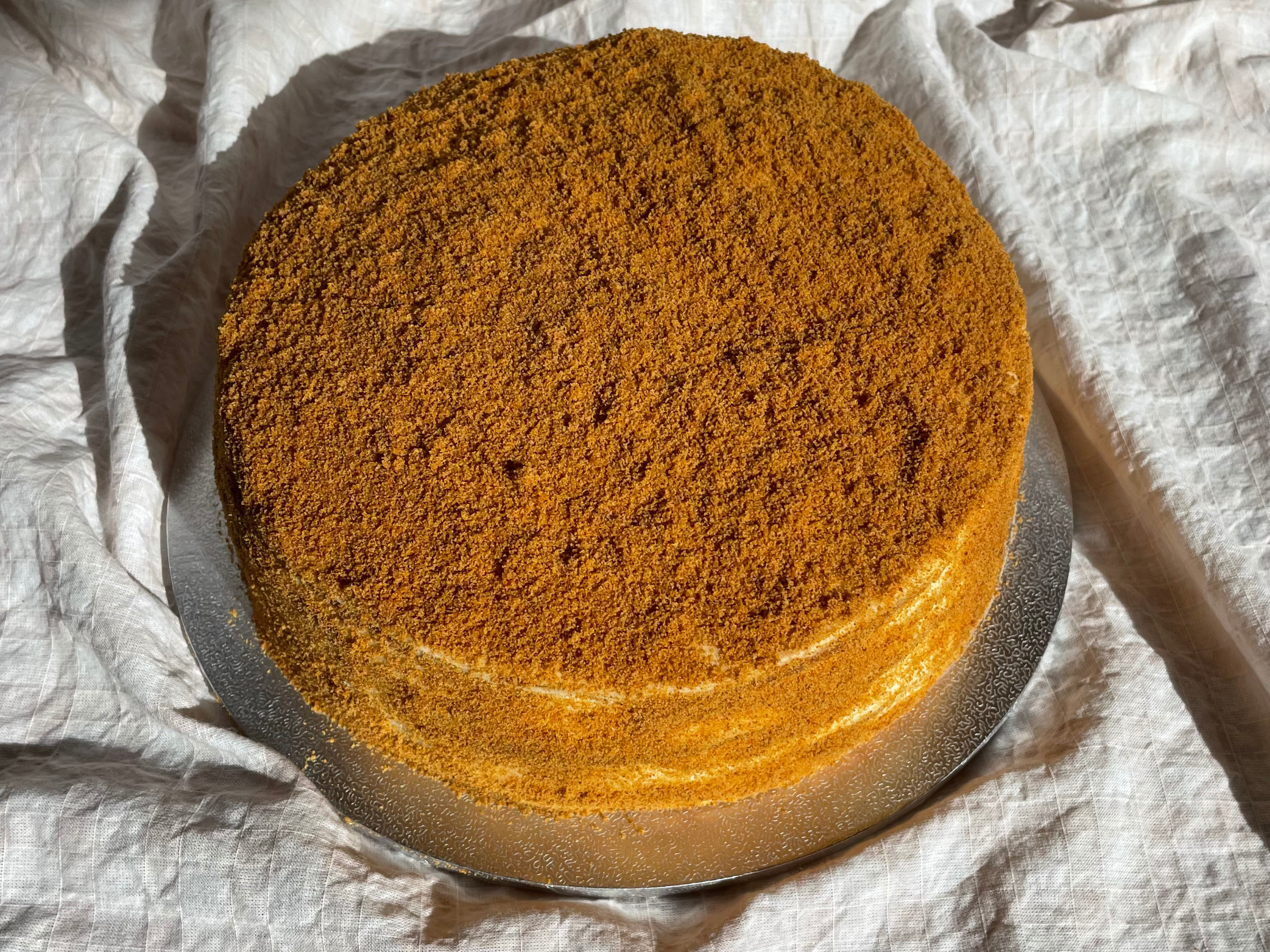 Golden honey layer cake covered in fine honeyed crumbs, sitting on a silver cake board.