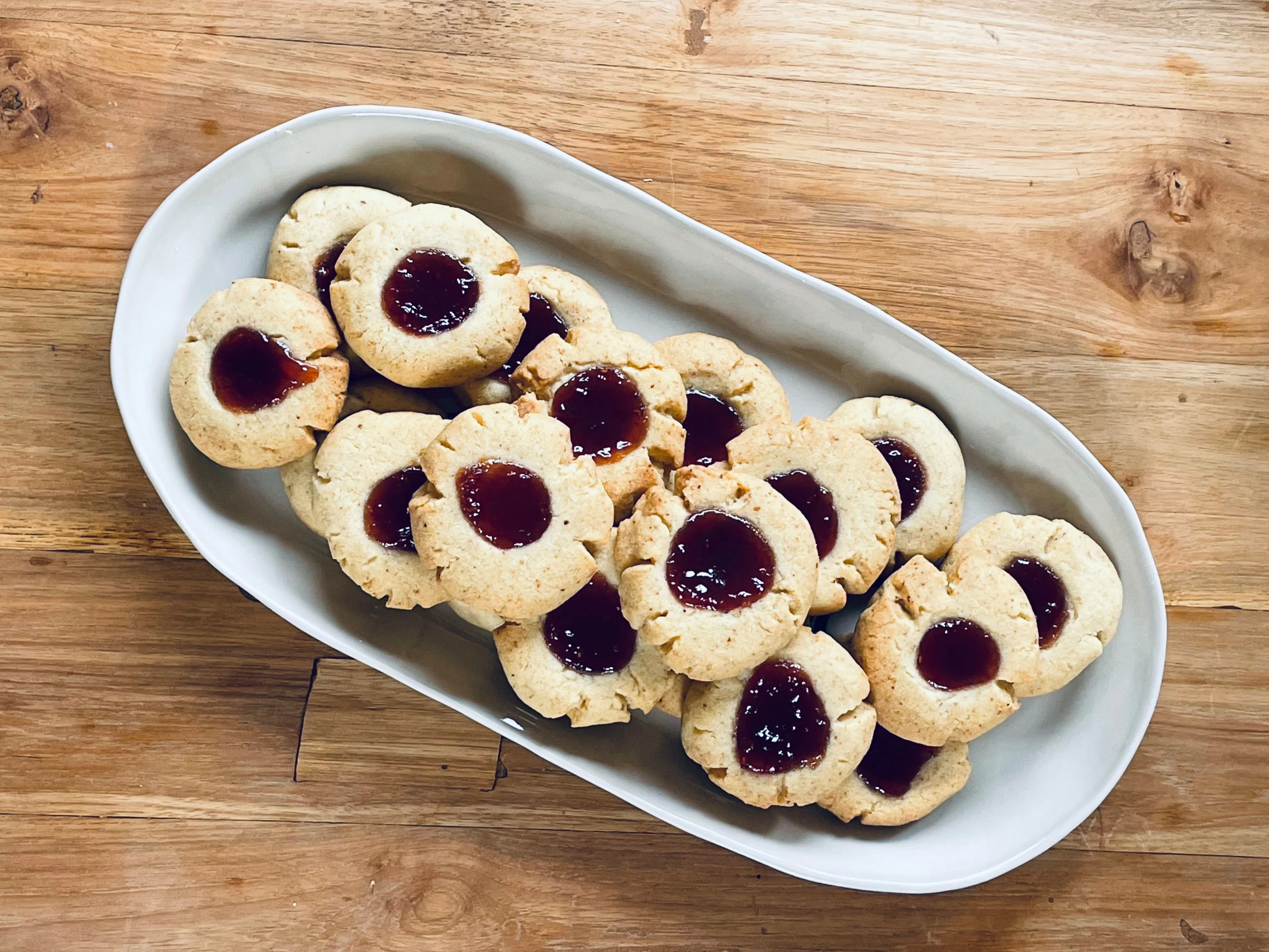 A white ceramic dish filled with golden brown butter jam drop biscuits, each with a centre of glossy raspberry jam, set on a rustic wooden table.